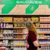 Prateleiras de supermercado com suplementos alimentares e produtos saudáveis, enquanto consumidora passa pelo corredor.